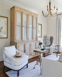 ASHLEY GILBREATH INTERIOR DESIGN: A simple sophisticated sitting area in the primary bedroom of a Southern Georgia lakeside home. When designing a space with a tv in the bedroom, Ashley loves to conceal the tv as much as possible. So an oversized, antique armoire cabinet usually does the trick. This bleached pine cabinet with a pleated cafe curtain grounds the space and is flanked by two unique arm chairs and iron bistro table.