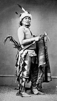 Dakota Indians: Portrait of Siha Han Ska (Long Foot) in Partial Native Dress with Headress and Ornaments and Holding Pipe Tomahawk - 1867