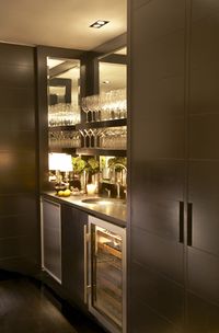 Sleek modern wet bar with espresso cabinetry. The bar area is flanked by cabinets, it features a mirrored interior and espresso stained floating shelves for glasses. A built-in mini fridge and glass fronted wine fridge sit below the counter. A black countertop is paired with an undermount bar sink and polished nickel faucet. Paul Davis - New York
