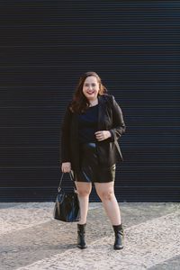 Look plus size usando blazer preto e saia de couro, bota de cano curto, bolsa bucket saco e batom vermelho