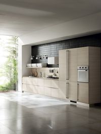 The white painted metal wall lamps with an exclusive design, positioned near the work area, make up a play of light and shade that offers warm, intimate atmospheres within the kitchen. The coordinated plinths and recessed grip profiles are made of aluminium, the sole material acting as a counterpoint to the Kuuki Elm decorative melamine that dominates the composition.