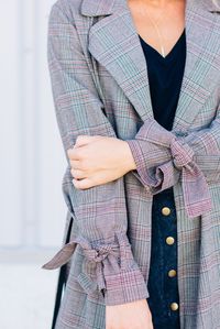 A woven lightweight trench coat featuring an allover Glenurquhart plaid print, notched collar, storm flaps, long sleeves with tie detail cuffs, and a removable self-tie sash.