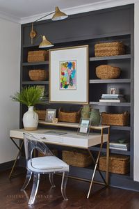 ASHLEY GILBREATH INTERIOR DESIGN: A cozy corner in a Southern lake home with floor to ceiling built-in dark grey shelves, brass swing arm light fixture, floating original framed artwork, and a lucite chair paired with a chic writing desk.