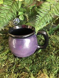 "This ceramic cauldron mug has been glazed with a high gloss purple shimmer glaze over my signature raven-black glaze on the body of the mug leaving the feet, handle, rim and interior of the mug in black. It has been fired to a temperature of 1940 degrees Fahrenheit so, it can take the heat and it is food safe! Enjoy your hot coffee, tea, or other hot or cold witch's brews from your own cauldron mug! It measures 4¼\" H x 3¼\" in diameter and holds 16 ounces. The three feet have a smooth glazed f