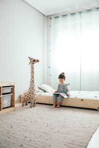 Niña leyendo en su habitación, sentada sobre la cama y acompañada de una jirafa de peluche gigante
