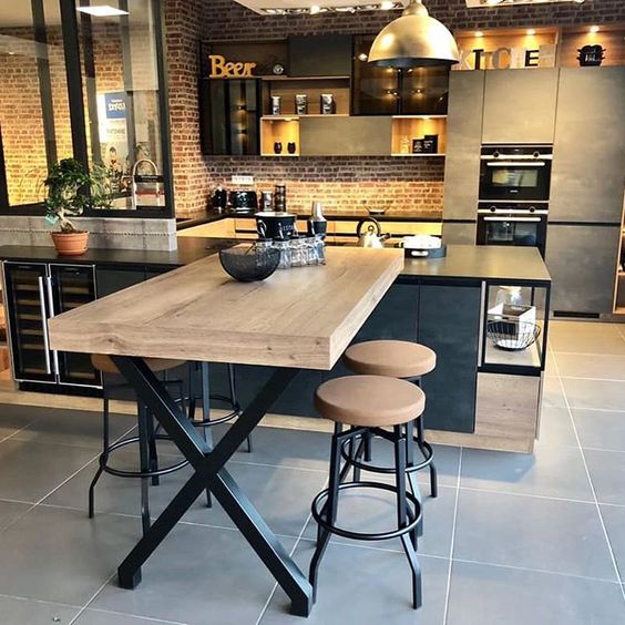 Une table industriel, un mur en brique, une cave à vin encastrée et des meubles acier et bois pour une cuisine noire et bois réussie