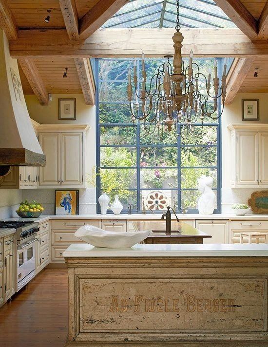 A stunning example of French Country mixed with modern farmhouse style in this rustic kitchen. Modern steel windows left unadorned and a wood ceiling mix with creamy painted cabinets and an antique work table. #farmhousestyle #Frenchkitchen #Frenchcountry #kitchendesign