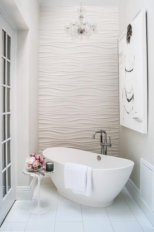 Romantic bathroom features an accent wall clad in wavy tiles alongside a crystal chandelier hanging over a corner freestanding tub and floor mount tub filler as well as a marble Saarinen Side Table.