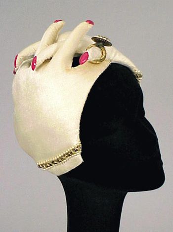 Bes-Ben clasping hands hat | Padded ivory velvet hands with applied red plastic fingernails, large pearl, and gilt-metal ring and pearl bracelets | Made in Chicago, United States.