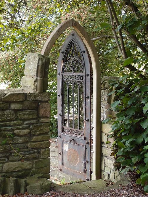 Garden gate ideas ad inspiration: gothic arch door flanked by stone walls in a lush green garden. #european #gardengate #gothic #gardendecor