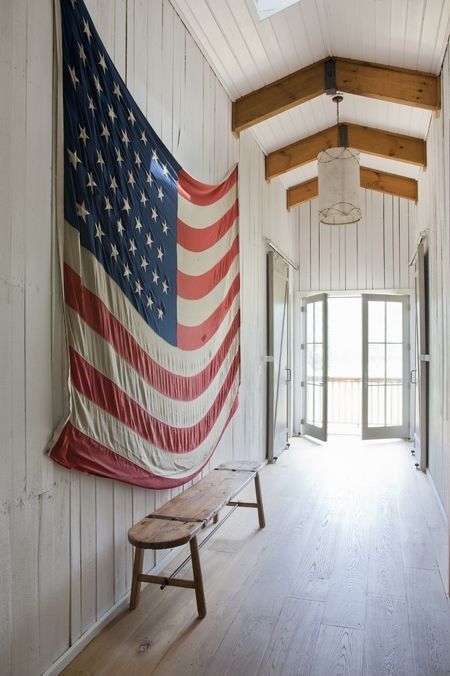 American flag hung on the wood paneled wall of a barn interior with beautiful country charm, a rustic bench, and skylight. #americana #patriotic #decor #july4