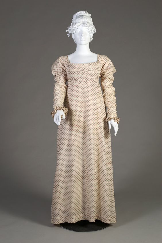 1808-12 White cotton day dress printed with red and blue floral rondels overall. The dress with scoop neck and high waist. A panel from the waistband flaps up over the bust, ties at the waist are pulled to the tightly pleated back. The short sleeves with sewn in fitted undersleeve with ruffled wrist. Silverman/Rodgers Collection, KSUM 1983.1.28