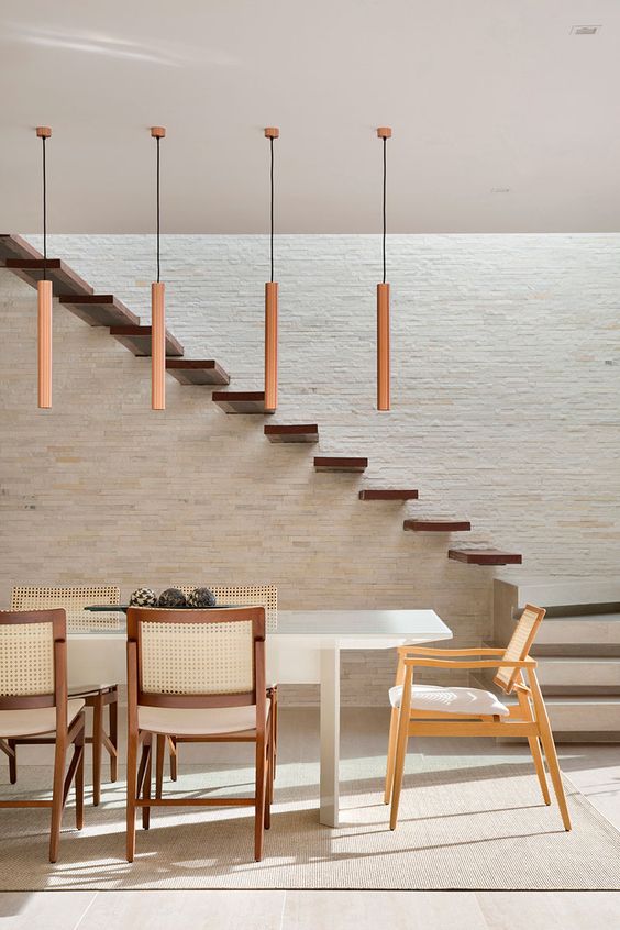 Sala de jantar moderna com mesa em laca branca, cadeiras em estilos diferentes, pendentes em cobre e parede texturizada com pedras naturais