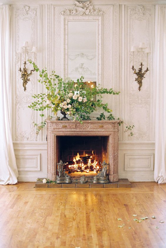 French fireplace in a Paris apartment Inspiration from Paris apartment interior design ideas - a charming living room with stone fireplace and paneled walls with ornate brass sconces and a magnificent floral arrangement.