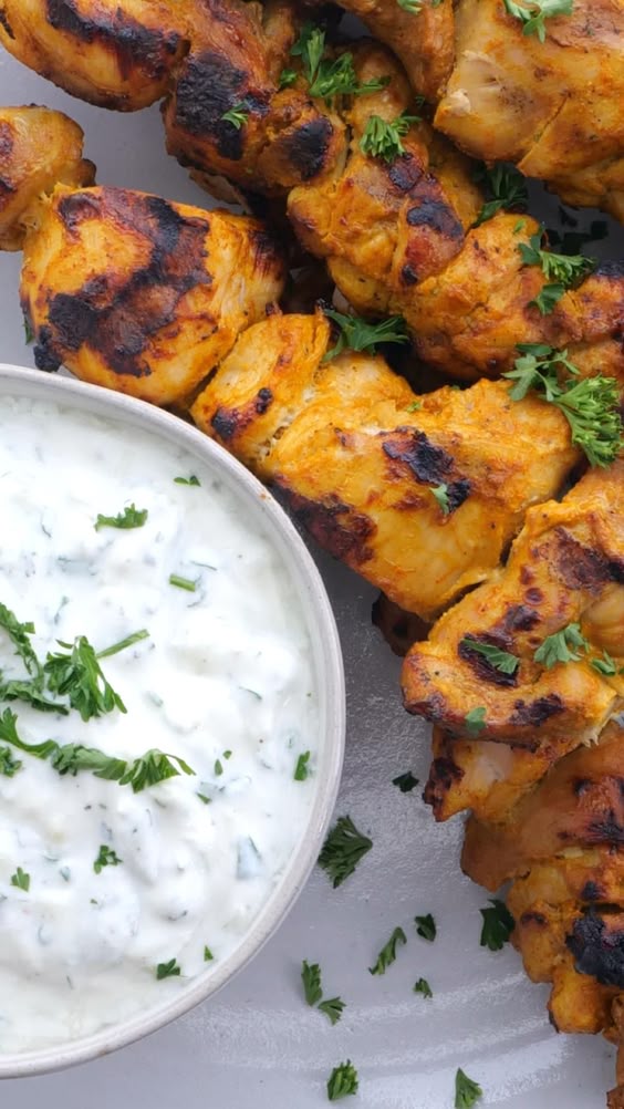 Video shows chicken marinade being made and chicken being cut up and put into marinade. Tzatziki sauce is made and whisked together. Chicken is removed from marinade and threaded onto skewers. Chicken is grilled and then served with tzatziki as a dipping sauce.