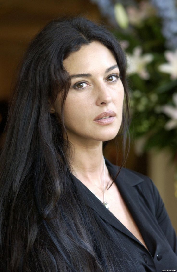 a woman with long black hair standing in front of flowers and looking at the camera