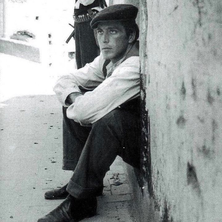 a man sitting on the side of a building next to another man wearing a hat