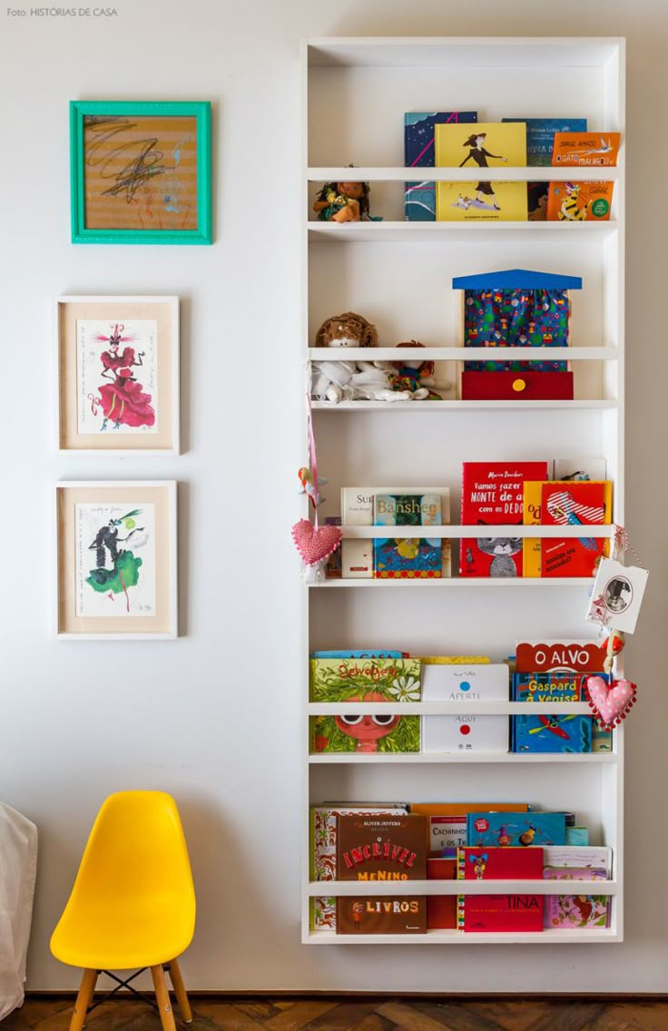 a white book shelf filled with books next to a yellow chair and pictures on the wall