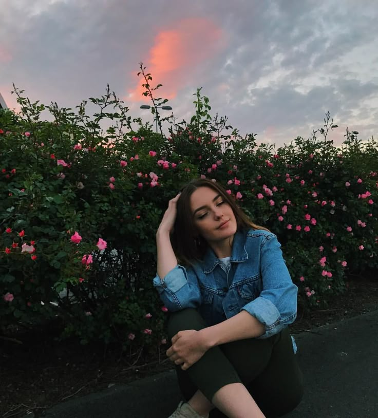 a woman sitting on the ground in front of some bushes with pink flowers at sunset