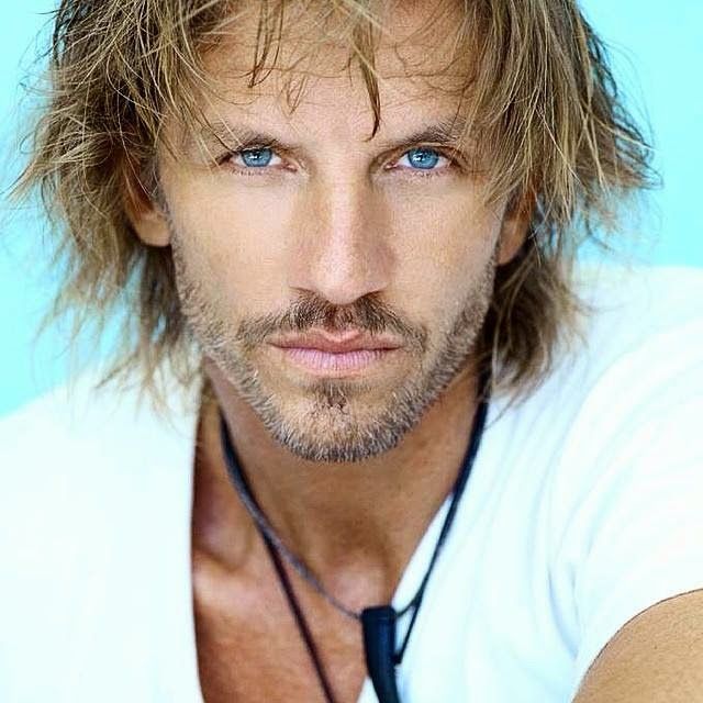 a man with long hair and blue eyes wearing a white shirt is posing for the camera