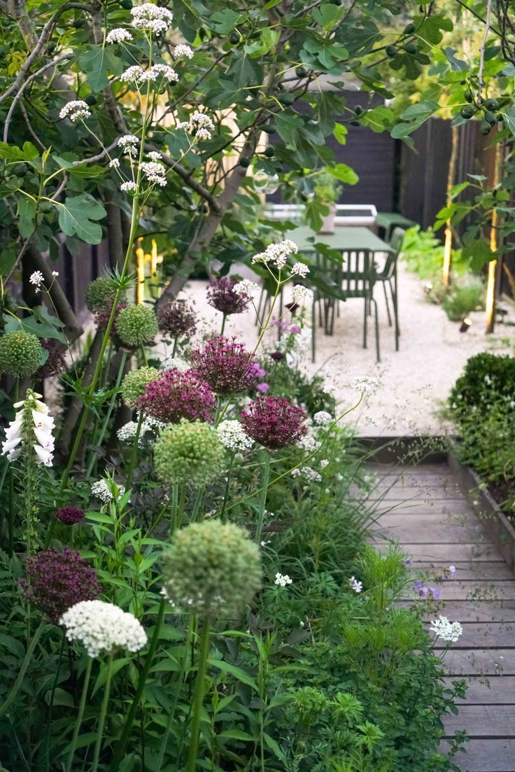 a garden with lots of plants and flowers in the foreground, next to a table and chairs