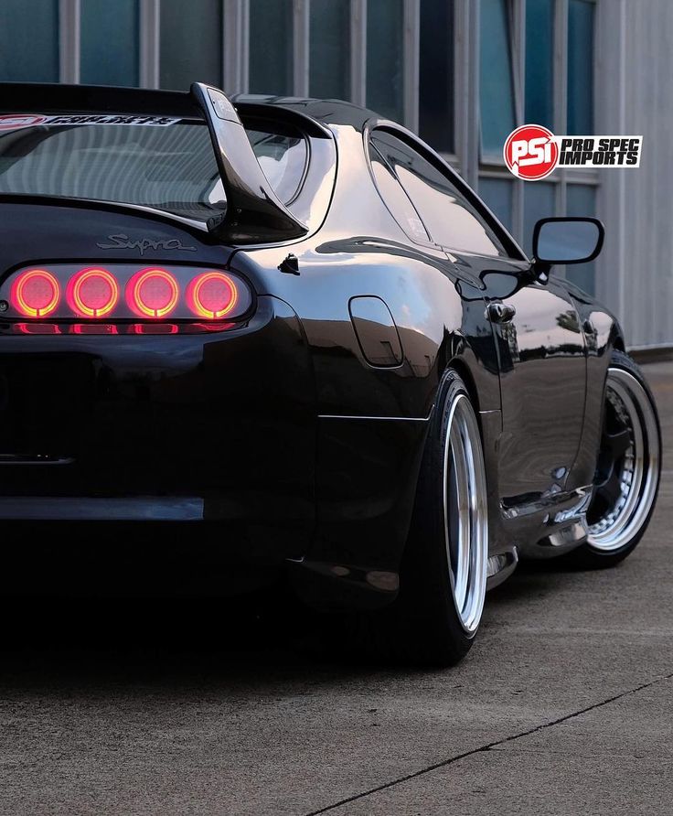 the rear end of a black sports car parked in front of a building with its lights on