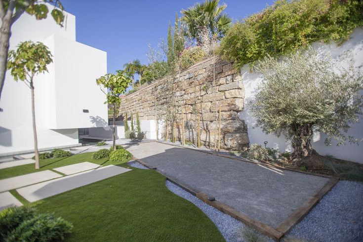 Un jardín contemporáneo y mediterráneo para una vivienda de estilo moderno. El proyecto consistió en decorar el jardín con complementos y recursos que se integrasen con la arquitectura. Rodeando la vivienda se diseñó un estanque semi-cubierto con cantos de acero que le aporta sonoridad y movimiento al exterior. También se jugó con distintos materiales en el pavimento, creando zonas de cerámica integradas en el césped y zonas de grava gris que crean un juego de caminos y un contraste con las form Contemporary Garden, Exterior, Sidewalk, Structures, Ponds, Crushed Stone, Trendy Tree, Driveways, Architecture