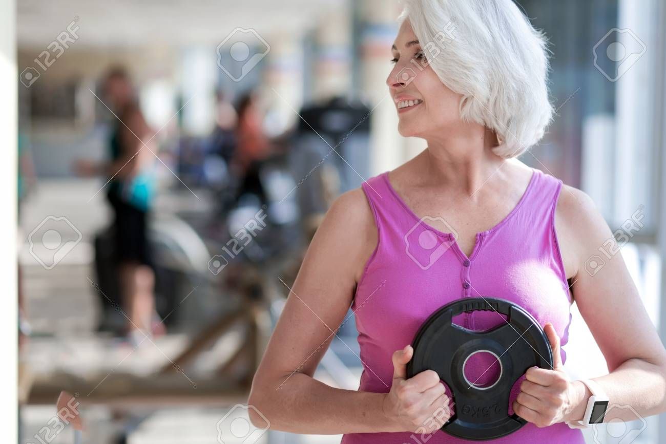 Happy fitness. Beautiful active woman turning head and smiling while exercising with plate in a gym....