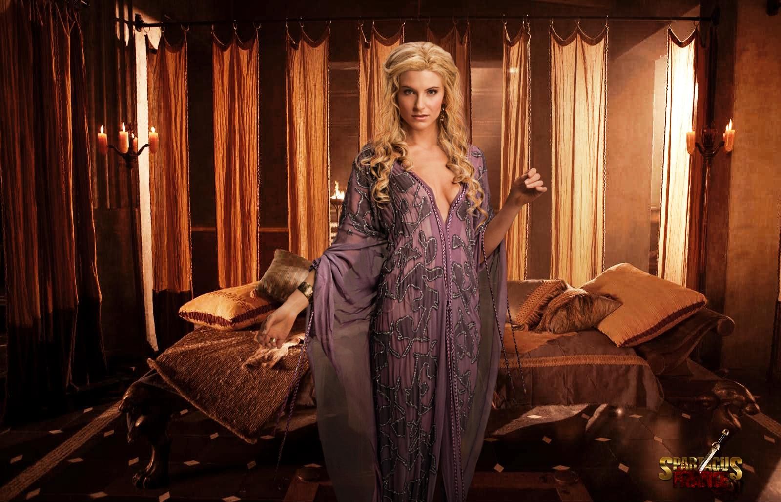 a woman standing in front of a bed wearing a purple dress and holding her hand up