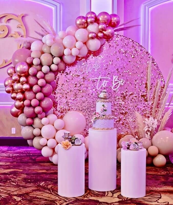Beautify your bridal shower party backdrop using scintillating décor accents to impart delightful vibes all around. Position a round metal arch covered with a pink sequin spandex cover and deck it up further using metallic rose gold balloons, pastel blush, dusty rose, and mocha latex balloons. Arrange some more balloons along with pampas grass beneath the stand while positioning white pedestal riser stands in front to make the overall décor theme look ethereal. Bridal Shower Backdrop, Bridal Shower Party, Metal Arch, Rose Gold Balloons, Pink Sequin, Pampas Grass, Backdrops For Parties, Riser, Latex Balloons