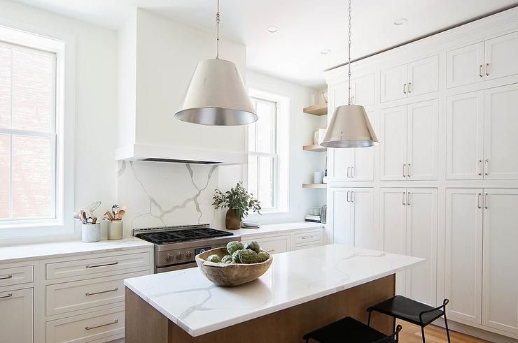Floor-to-ceiling white shaker cabinets fitted with nickel pulls are positioned facing a brown stained island seating low back black metal counter stools at a statuary quartz countertop lit by two nickel pendants. #whiteshakercabinets