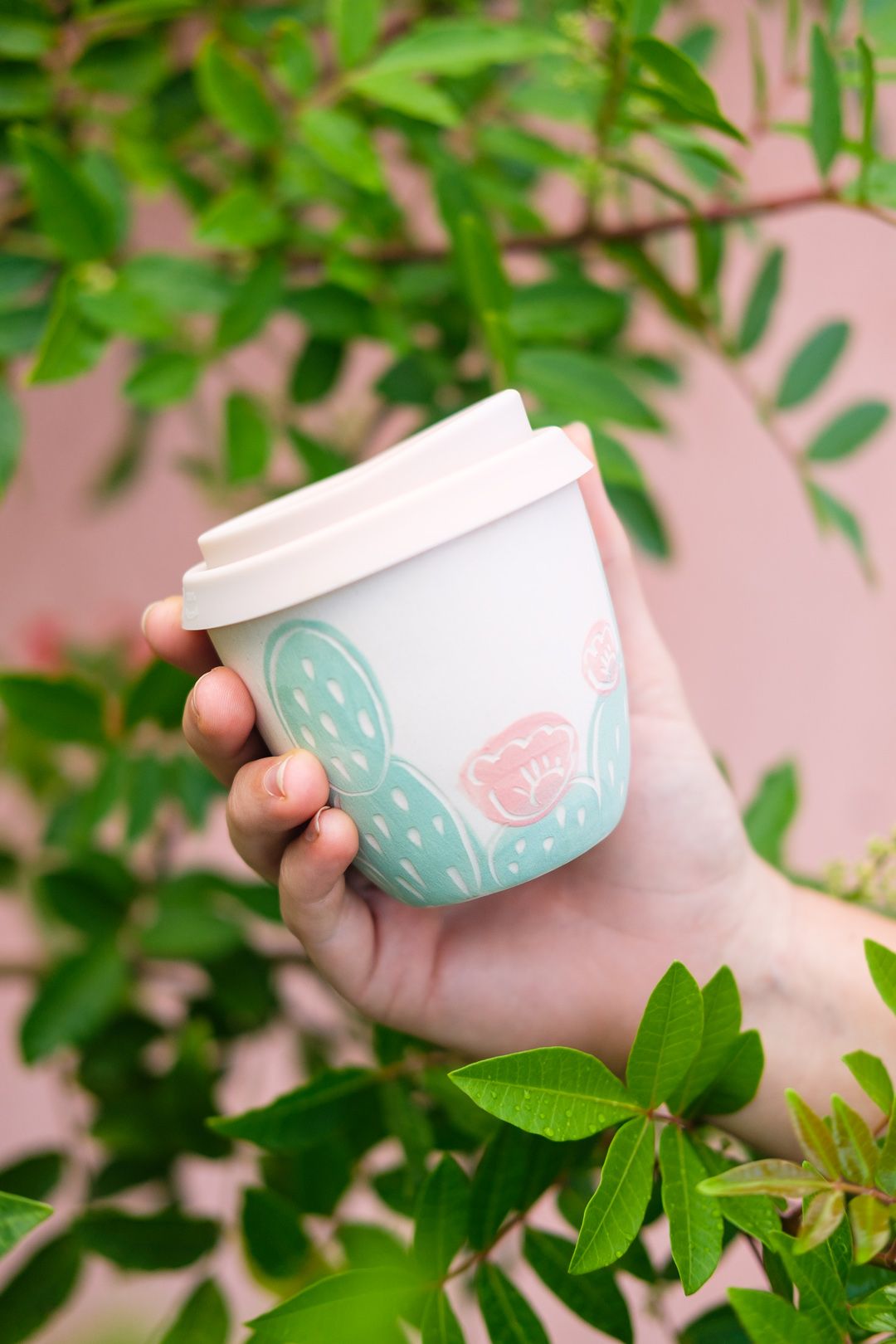 A Hand Holding A Coffee Cup In Front Of Some Green Leaves On A Pink Wall a-hand-holding-a-coffee-cup-in-front-of-some-green-leaves-on-a-pink-wall